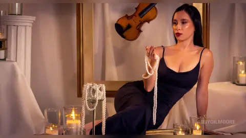 Elegant woman in a black evening dress holding a pearl necklace in a sophisticated, moody candlelit room.
