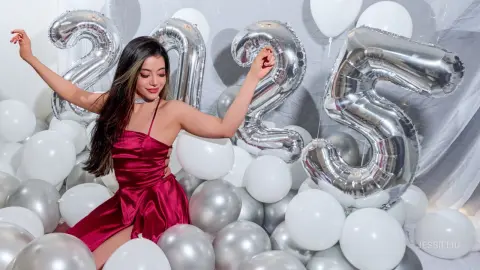 JessieLiu poses behind silver and white balloons wearing a red satin slip dress.