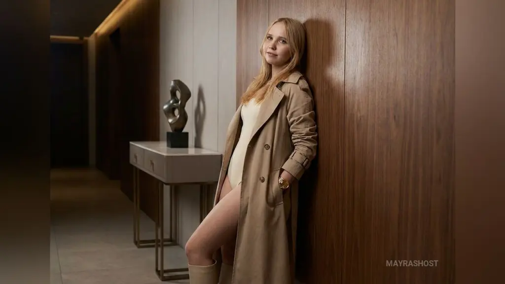 Blonde woman in a cream satin dress sitting by a bouquet of roses and glowing candles in a classic room.