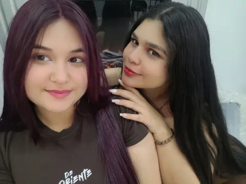 Two smiling young women with long dark hair posing closely in a cozy indoor setting.