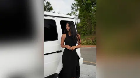 Smiling woman in a chic black sleeveless dress standing next to a white luxury van in a bright outdoor setting.