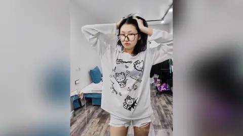 TishaOyervides wears a white long-sleeve shirt with cartoon graphics and white denim shorts while holding her black hair up with both hands.