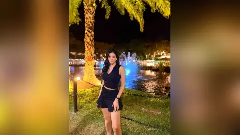 Elegant woman in a black sleeveless outfit standing by a lake with a fountain at night.