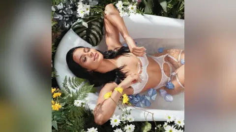 Relaxed woman in white swimwear lounging in a white bathtub surrounded by lush green ferns and white flowers.