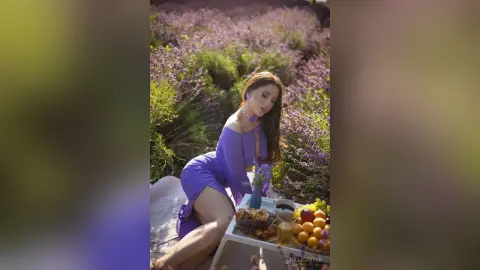 Young woman in a purple dress sitting in a lavender field with a summer picnic spread.