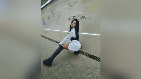 KymSkyler poses in a white shirt and short white skirt with black boots, leaning against a light-colored wall with graffiti in a subway setting.