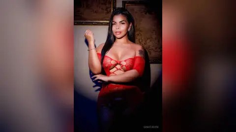 Glamorous woman in a red lace bodysuit posing in a dimly lit room with a moody, elegant atmosphere.