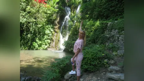 Blonde woman in pink loungewear standing by a lush jungle waterfall in a serene natural setting.