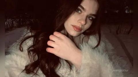 Young woman with long wavy hair wearing a cozy white faux fur coat in a snowy outdoor setting at night.