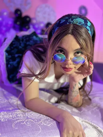 maaeve_elf — Young woman in retro blue sunglasses and headband lying on a bed in dreamy purple lighting.