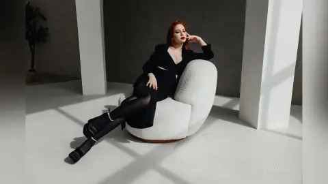 Elegant redhead in a chic black outfit relaxing on a white bouclé chair in a minimalist studio setting.