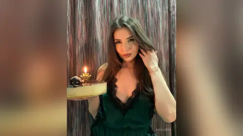 Beautiful woman in a green lace dress holding a birthday cake with a lit candle in a cozy indoor setting.