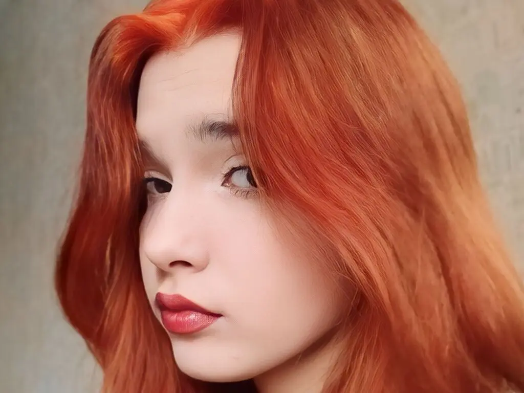 Close-up portrait of a young woman with vibrant long red hair and natural makeup looking softly at the camera.