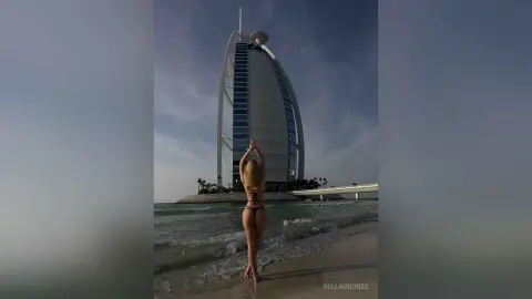 BellaBronze stands naked on a beach facing a large, sail-shaped building, showing her bare back and buttocks while raising her arms above her head.