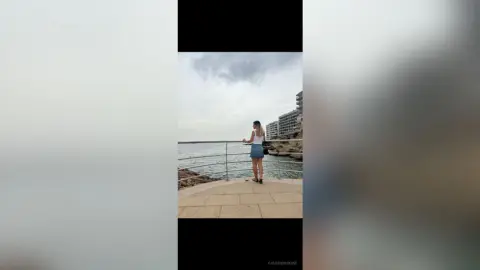 Young woman in a white tank top and denim skirt standing on a coastal walkway overlooking the ocean under a cloudy sky.