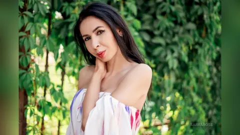 Elegant woman in a white off-the-shoulder floral dress posing in a lush green garden setting.