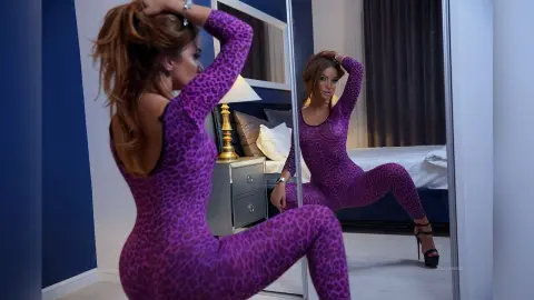 FelishaRose poses in a purple leopard print bodysuit while looking into a mirror in a bedroom setting.