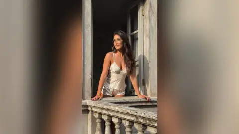 Young woman in white satin loungewear posing on a sunlit stone balcony during golden hour.
