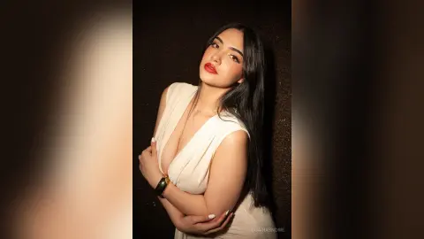 Elegant woman in a white sleeveless blouse posing with red lipstick against a dark studio background.
