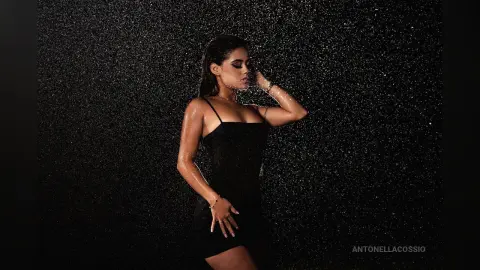 Glamorous woman in a black cocktail dress posing under falling water against a dark background.
