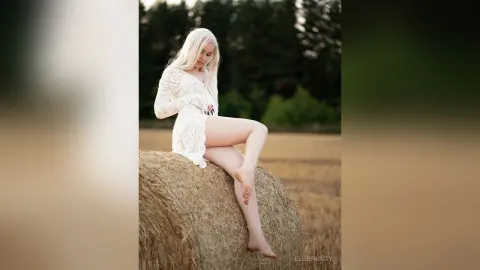 Blonde woman in a white lace dress sitting on a hay bale in a golden field during sunset.