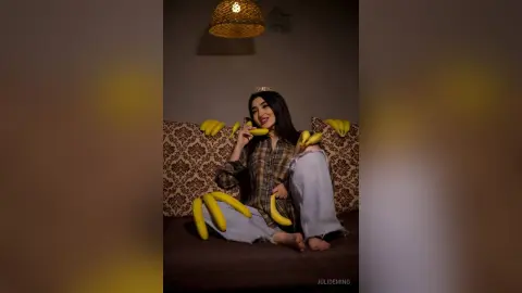 JuliDeming sits on a patterned couch wearing a plaid shirt and light jeans, surrounded by many yellow bananas.