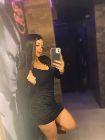 Young woman in a sleek black mini dress taking a mirror selfie in a moody, dimly lit lounge setting.