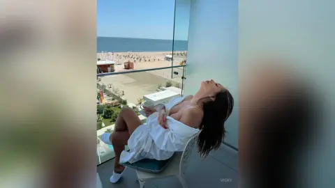 Relaxed woman in a white robe lounging on a balcony overlooking a sunny beach and ocean view.