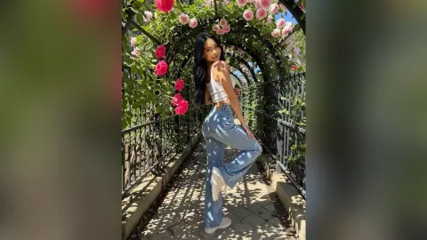 Smiling young woman in white tank top and blue jeans posing in a sunny rose garden walkway.