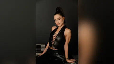 Bella Poarch in a black lace-up leather vest and trousers posing confidently against a dark studio background.