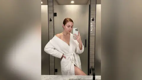 Young woman in a white waffle robe taking a mirror selfie in a modern minimalist bathroom.