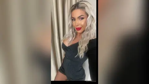 KateJordann poses in a dark grey off-the-shoulder top, showing her long blonde wavy hair and red lipstick against a neutral curtain background.