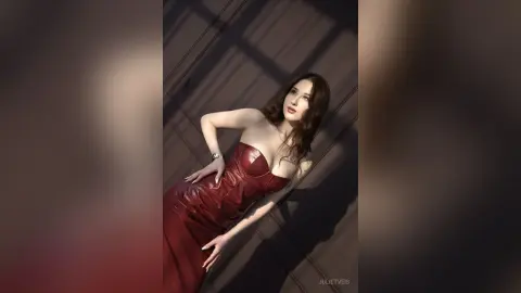 Elegant woman in a red satin evening gown posing in dramatic sunlight against a dark architectural background.