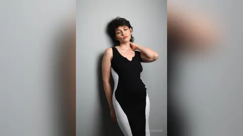 IveteDurand poses against a grey wall wearing a tight black and white dress, touching her neck with one hand.