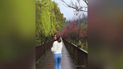 Young woman in a white fuzzy sweater and blue jeans walking on a wooden bridge through a lush green forest.
