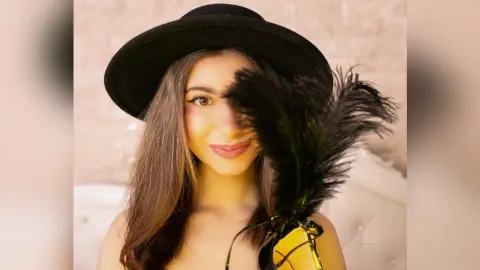 Stylish woman in a black wide-brimmed hat holding black feathers in a bright, elegant setting.