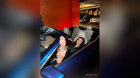Young woman in a black striped top relaxing barefoot in a blue convertible car in a dimly lit parking garage.