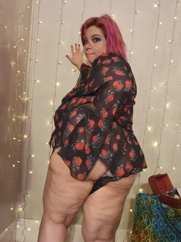 Curvy woman with pink hair in a black floral sheer blouse posing against fairy lights in a cozy room.