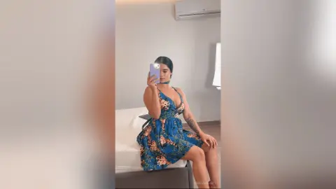 Young woman in a blue floral sundress taking a mirror selfie in a bright, modern bedroom.