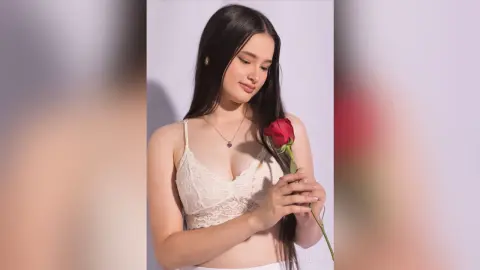 Young woman in a white lace camisole holding a red rose against a plain background with a romantic mood.