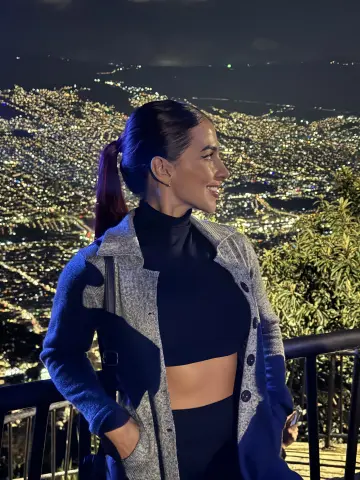 Smiling woman in a black turtleneck and grey cardigan overlooking a sparkling city skyline at night.