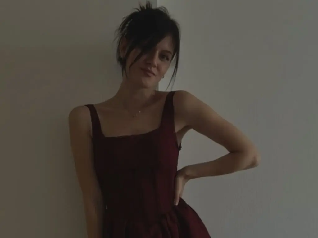 Young woman in a dark red sleeveless dress posing against a neutral wall in soft indoor lighting.