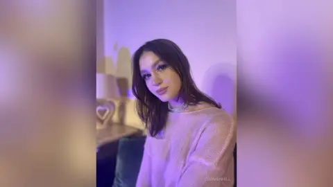 Young woman in a pink knit sweater posing in soft purple ambient lighting for a dreamy, moody portrait.