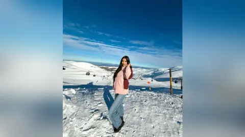 Aleja stands in a snowy mountain landscape wearing a pink long-sleeved top and light blue jeans.
