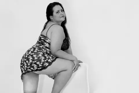 Curvy woman in floral lace lingerie sitting on a white chair against a plain background in a black and white portrait.