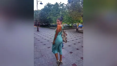SandraMidnight stands on a stone walkway wearing a light blue sleeveless dress and carrying a patterned handbag.