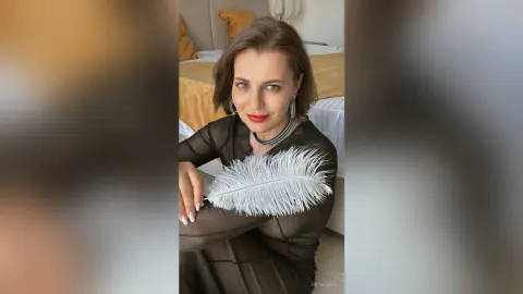 Elegant woman in a black long-sleeve dress with white feather details posing in a bright bedroom.