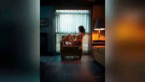 Nude woman sitting in a chair by a window in a moody, dimly lit room with warm lamp lighting.