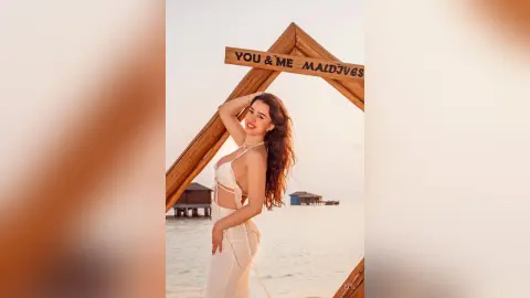 Elie poses on a beach wearing a white string bikini, standing under a wooden frame with the words 'YOU & ME MALDIVES' behind her.