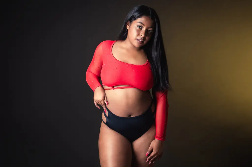 Smiling curvy woman in a red long-sleeve top and black shorts posing on a chair in a studio setting.
